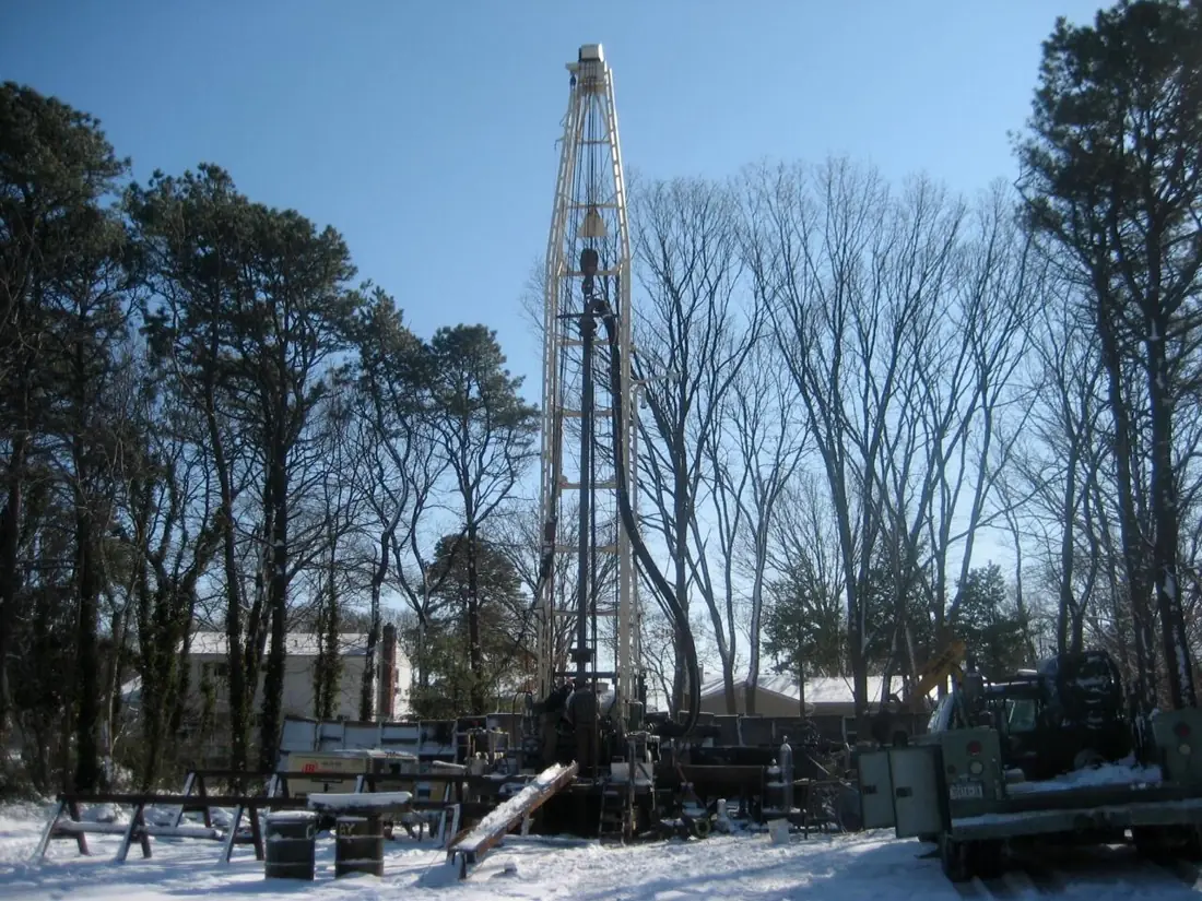 A tall drilling rig and support trucks at a well site among bare winter trees.